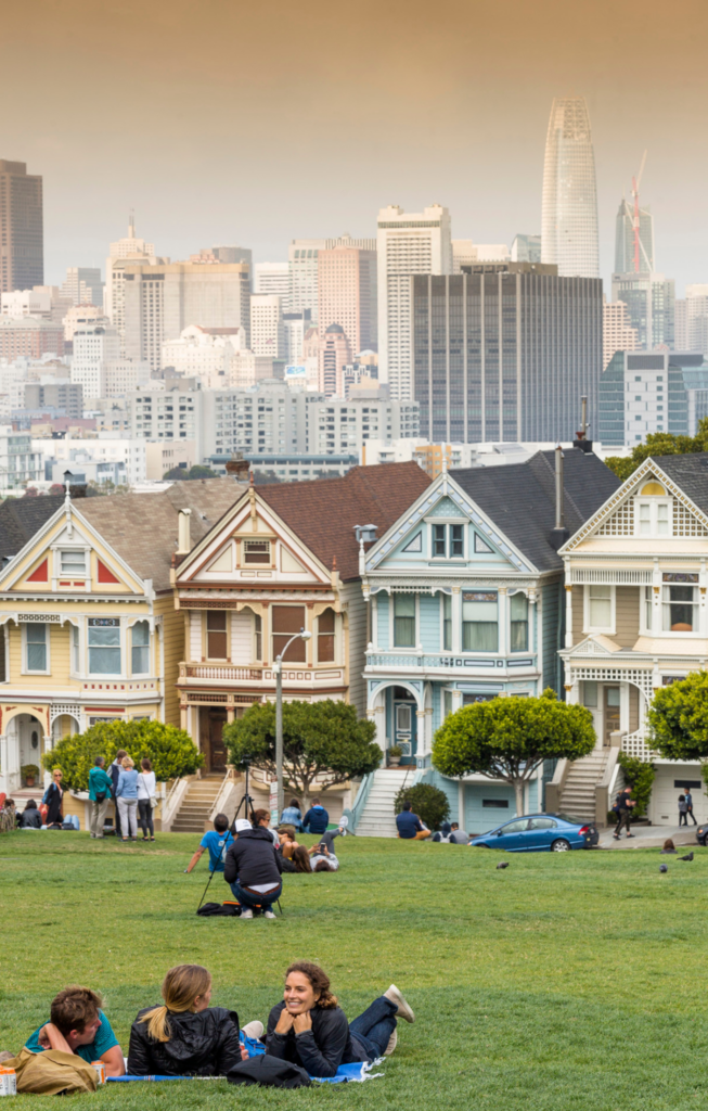 the four colorful homes known as the painted ladies in the middle ground with the park and people lounging in the foreground and in the background are the skyscrapers of san francsisco